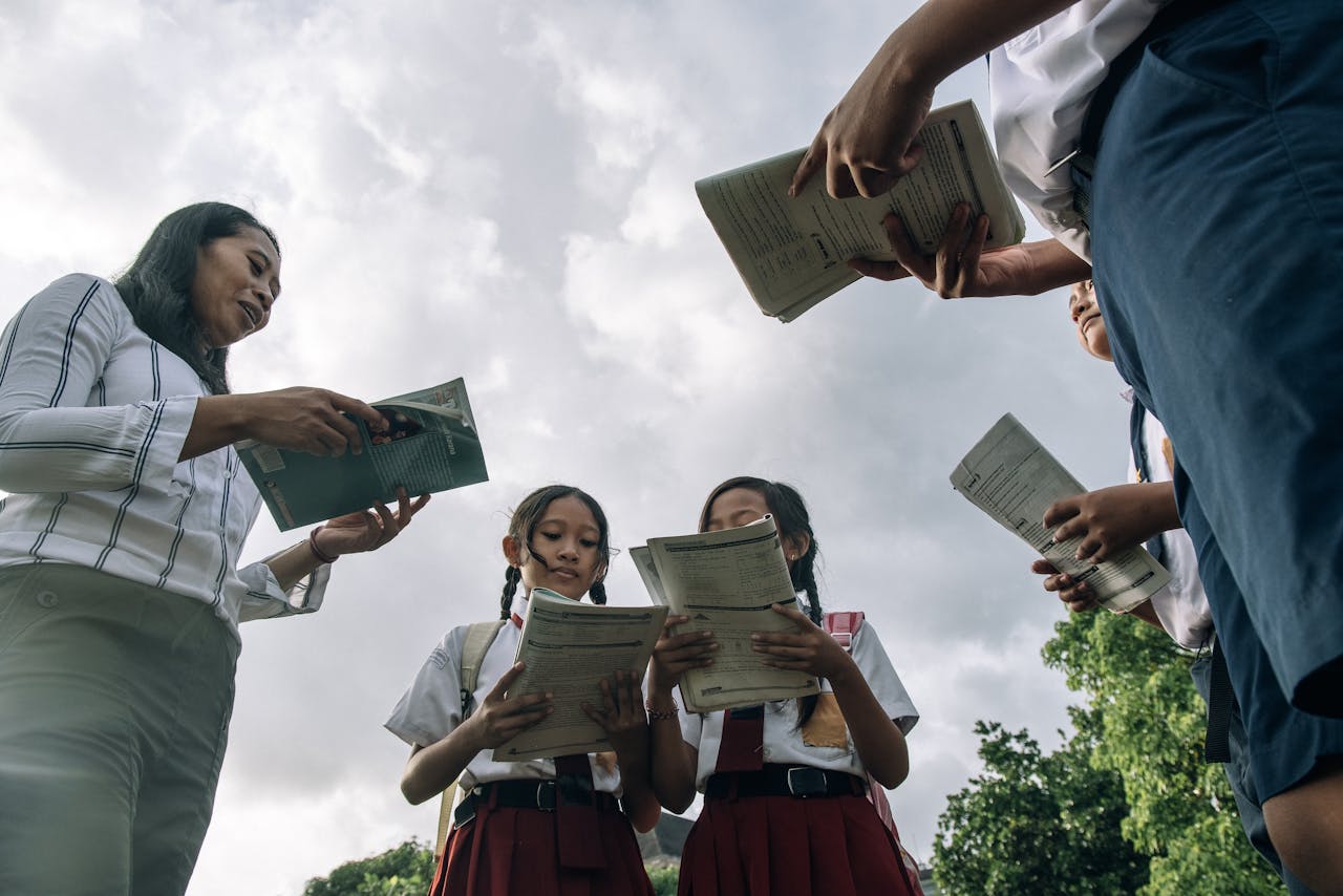 Students and teacher in school uniforms reading books outdoors under a cloudy sky.